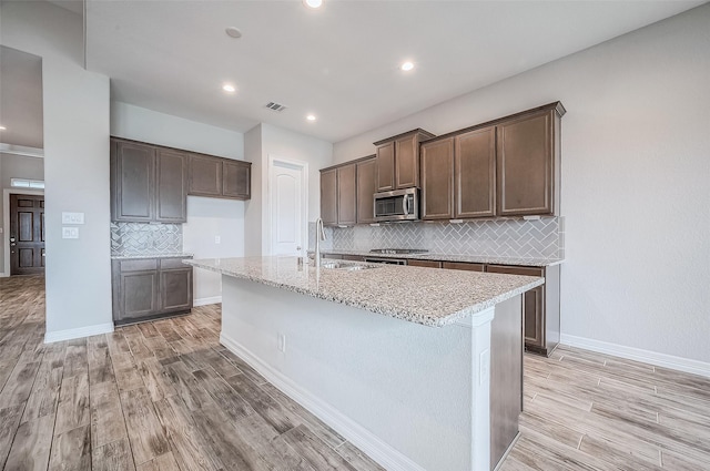 kitchen with light wood-type flooring, stainless steel appliances, backsplash, and an island with sink