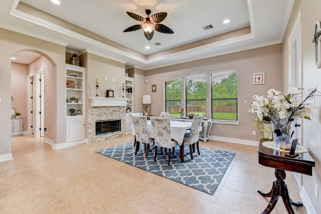 dining space with a stone fireplace, built in shelves, ornamental molding, ceiling fan, and a raised ceiling