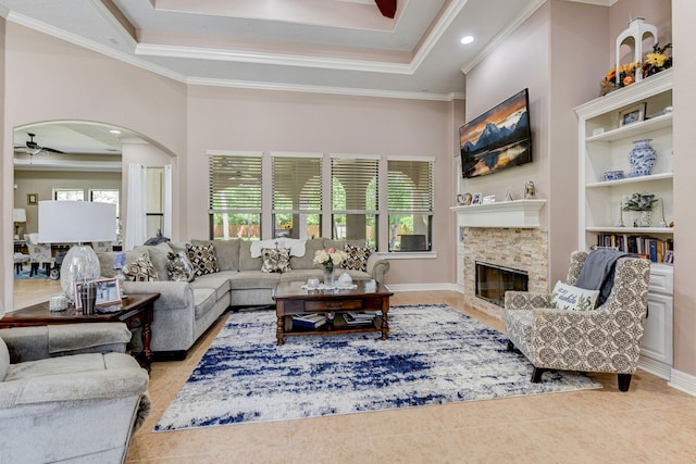 tiled living room featuring a stone fireplace, ornamental molding, ceiling fan, and a tray ceiling