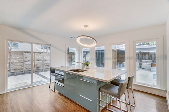 kitchen with decorative light fixtures, an island with sink, sink, stainless steel oven, and light hardwood / wood-style floors