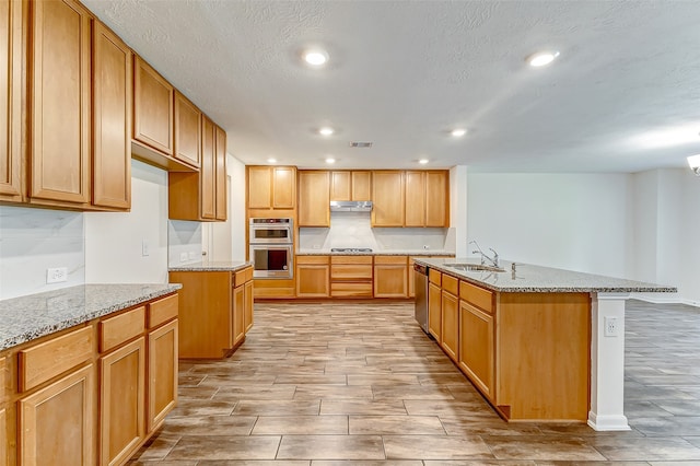 kitchen featuring appliances with stainless steel finishes, a textured ceiling, light stone countertops, an island with sink, and light hardwood / wood-style flooring