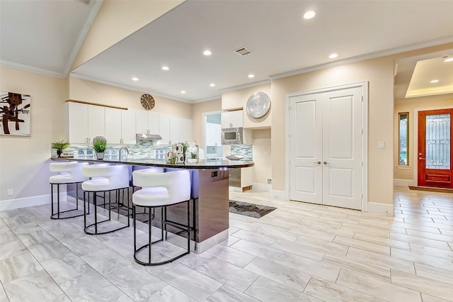 kitchen with tasteful backsplash, ornamental molding, a kitchen breakfast bar, white cabinets, and stainless steel microwave