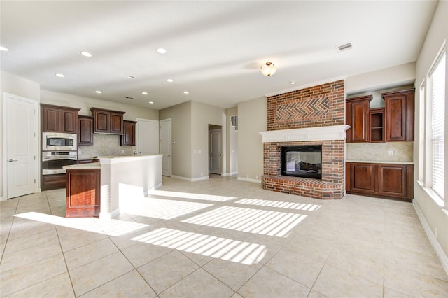 kitchen with appliances with stainless steel finishes, backsplash, a center island with sink, a fireplace, and light tile patterned flooring