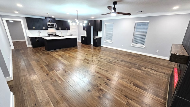 kitchen with dark wood-type flooring, an island with sink, ceiling fan, crown molding, and appliances with stainless steel finishes