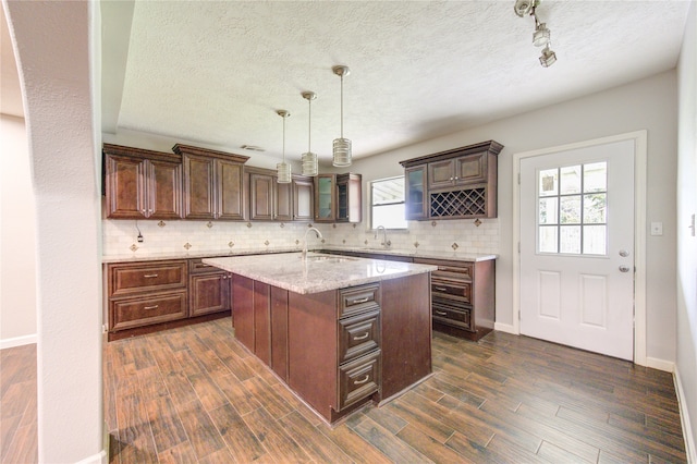 kitchen featuring dark hardwood / wood-style flooring, light stone counters, tasteful backsplash, an island with sink, and decorative light fixtures