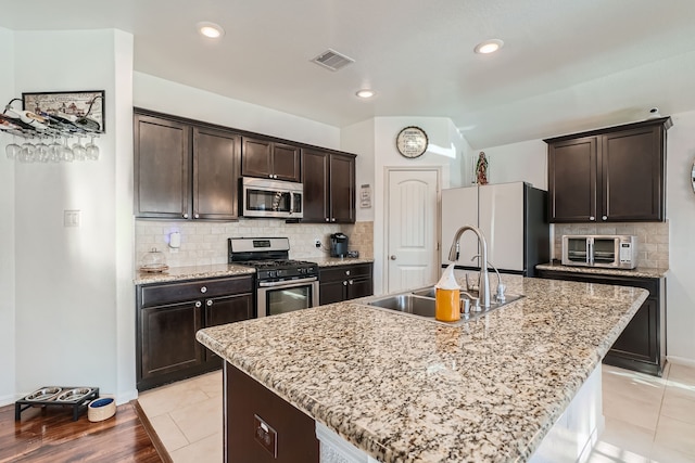 kitchen featuring an island with sink, sink, light stone counters, and appliances with stainless steel finishes