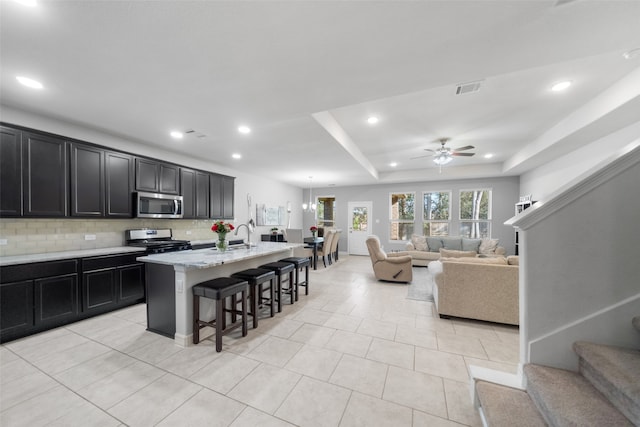 kitchen with backsplash, appliances with stainless steel finishes, a kitchen bar, an island with sink, and ceiling fan