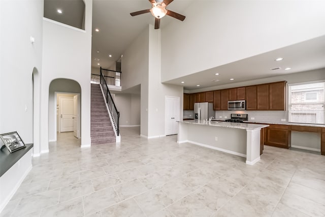 kitchen featuring stainless steel appliances, a center island with sink, light stone counters, ceiling fan, and a towering ceiling