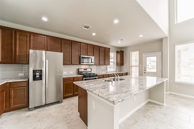 kitchen with stainless steel appliances, a center island with sink, sink, light stone countertops, and decorative backsplash