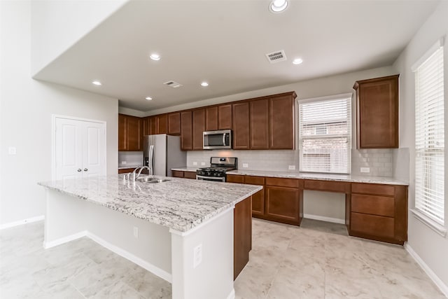 kitchen featuring stainless steel appliances, a center island with sink, sink, light stone counters, and tasteful backsplash