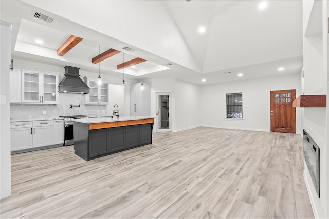 kitchen featuring a center island with sink, custom exhaust hood, stainless steel gas stove, white cabinetry, and decorative light fixtures