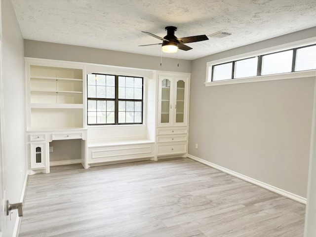 empty room with built in desk, ceiling fan, plenty of natural light, and light hardwood / wood-style flooring