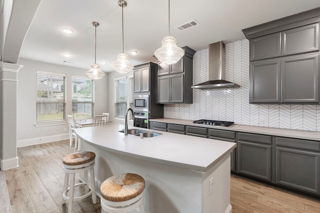 kitchen featuring light hardwood / wood-style floors, a kitchen island with sink, wall chimney range hood, appliances with stainless steel finishes, and decorative light fixtures