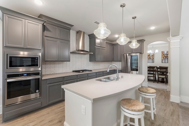 kitchen with stainless steel appliances, decorative light fixtures, sink, wall chimney exhaust hood, and light hardwood / wood-style flooring