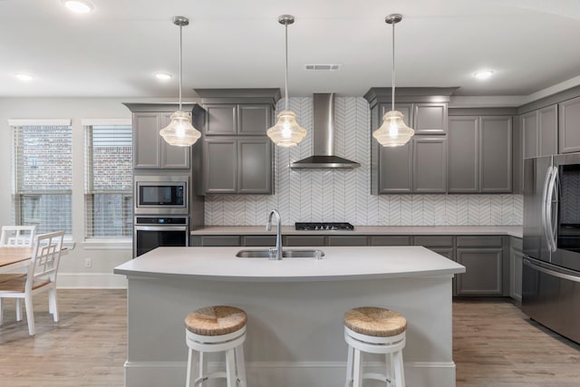 kitchen featuring wall chimney range hood, appliances with stainless steel finishes, pendant lighting, sink, and a kitchen island with sink