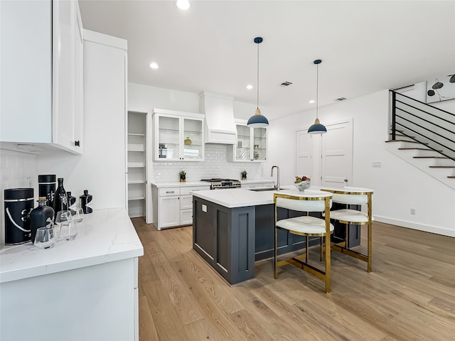 kitchen with stainless steel stove, premium range hood, an island with sink, white cabinets, and light wood-type flooring