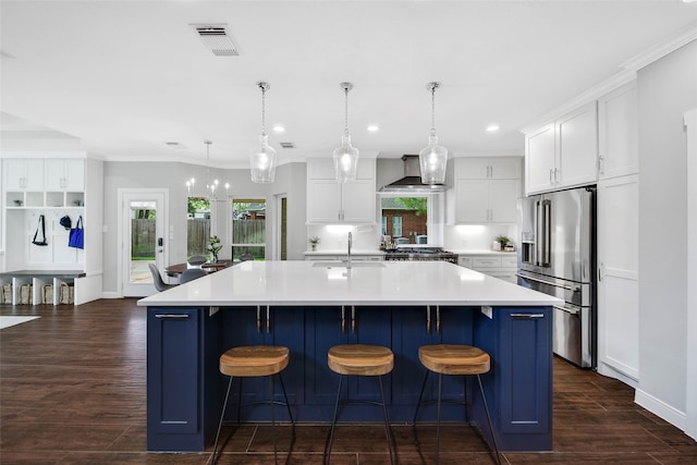 kitchen with a large island with sink, high end refrigerator, wall chimney range hood, decorative light fixtures, and white cabinetry