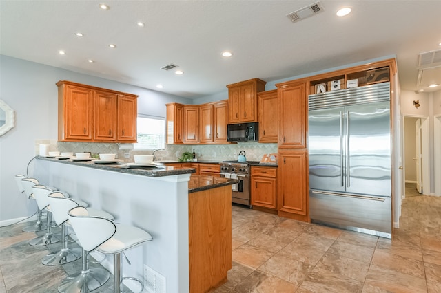 kitchen with kitchen peninsula, backsplash, dark stone counters, a breakfast bar, and premium appliances