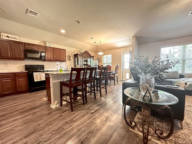 kitchen with dark wood-type flooring, black appliances, a kitchen island with sink, light stone countertops, and decorative light fixtures