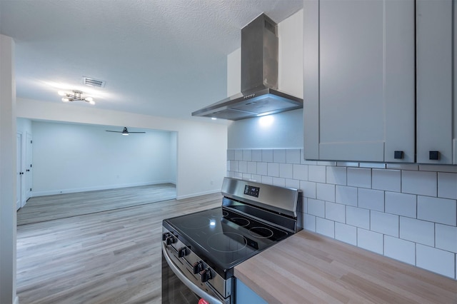 kitchen with light wood-type flooring, backsplash, a textured ceiling, wall chimney range hood, and stainless steel range oven