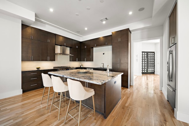 kitchen with an island with sink, stainless steel fridge, light stone countertops, dark brown cabinets, and wall chimney range hood