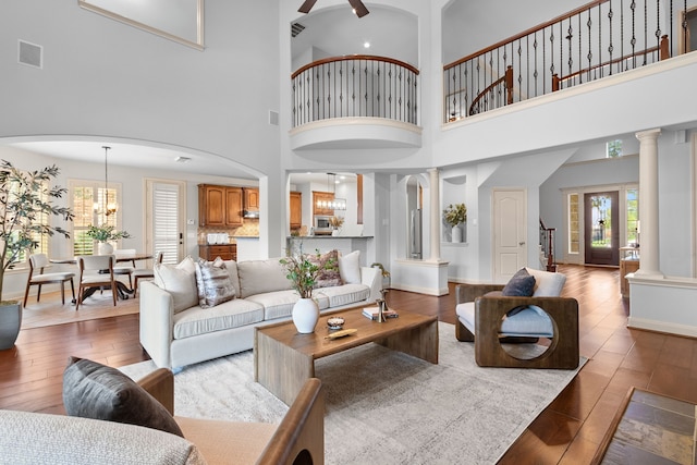 living room featuring a towering ceiling, ceiling fan with notable chandelier, ornate columns, and light hardwood / wood-style flooring