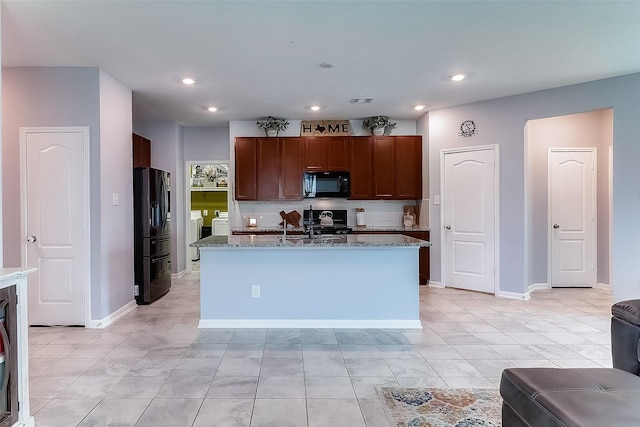 kitchen featuring stainless steel fridge, stove, washer and clothes dryer, a center island with sink, and dark stone countertops