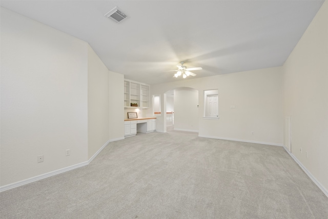 unfurnished living room featuring built in shelves, light colored carpet, built in desk, and ceiling fan