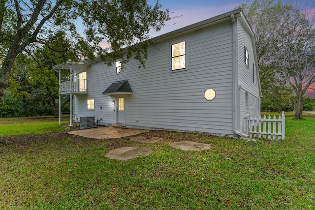 back house at dusk with a balcony, central air condition unit, a lawn, and a patio