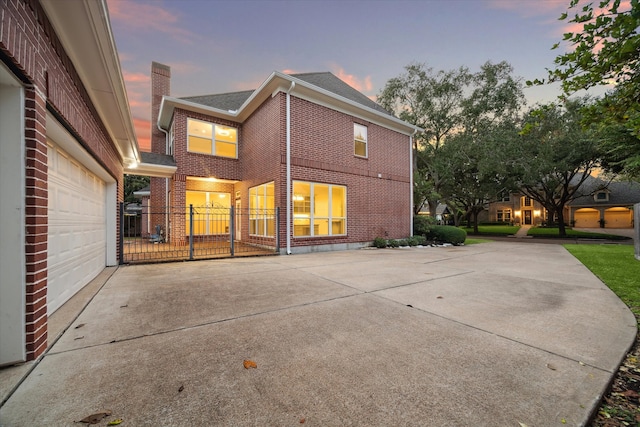 back of house at dusk with a garage, concrete driveway, brick siding, and a chimney