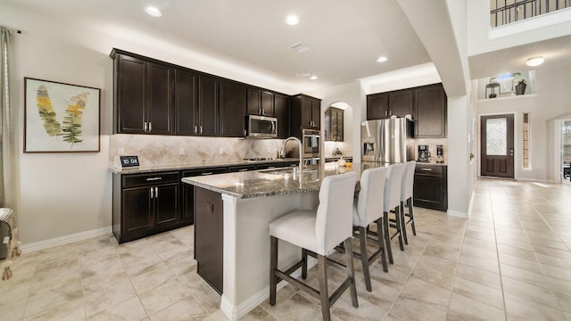 kitchen featuring dark brown cabinetry, stone counters, sink, stainless steel appliances, and a center island with sink