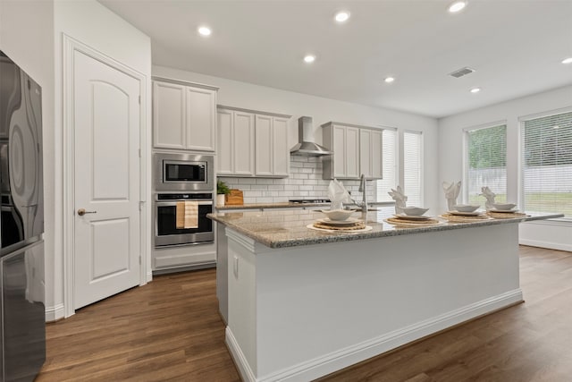 kitchen featuring light stone countertops, dark wood-type flooring, wall chimney range hood, white cabinets, and an island with sink