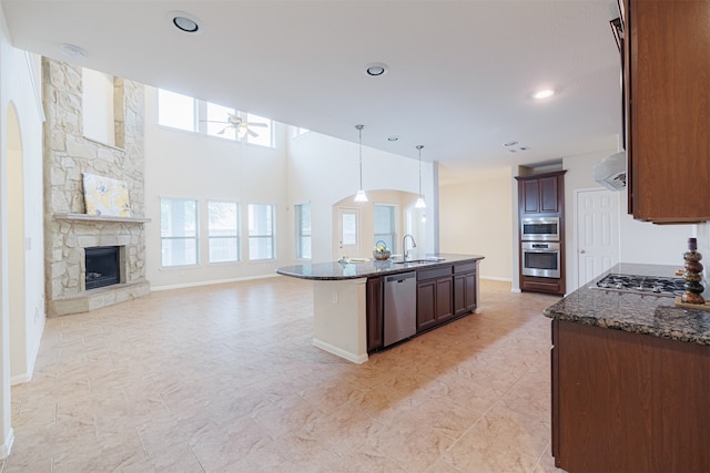 kitchen featuring stainless steel appliances, dark stone countertops, an island with sink, a stone fireplace, and range hood