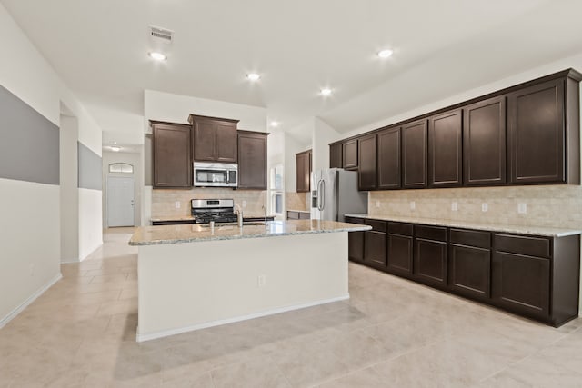 kitchen featuring stainless steel appliances, light stone countertops, backsplash, an island with sink, and dark brown cabinets