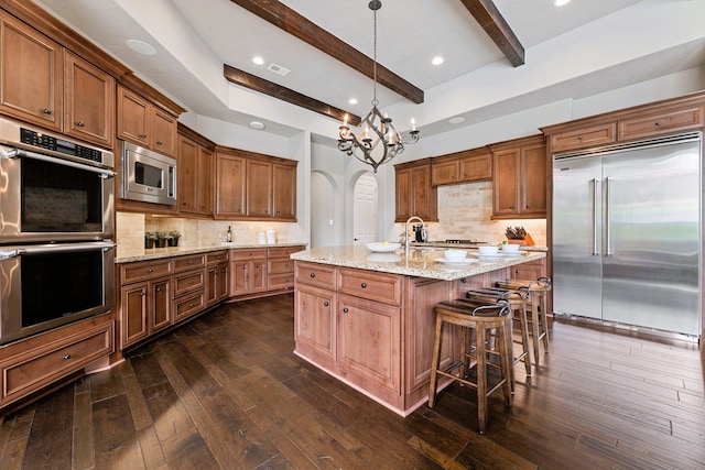 kitchen featuring dark hardwood / wood-style flooring, tasteful backsplash, built in appliances, a center island with sink, and beamed ceiling