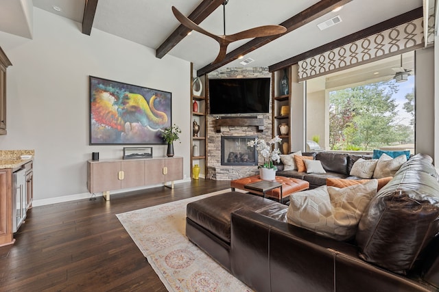 living room with built in shelves, ceiling fan, dark wood-type flooring, a stone fireplace, and beamed ceiling