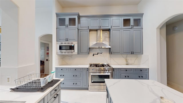4009 Ravencrest Ct, Pearland, TX 77584 - kitchen featuring wall chimney exhaust hood, appliances with stainless steel finishes, gray cabinetry, arched walkways, and backsplash