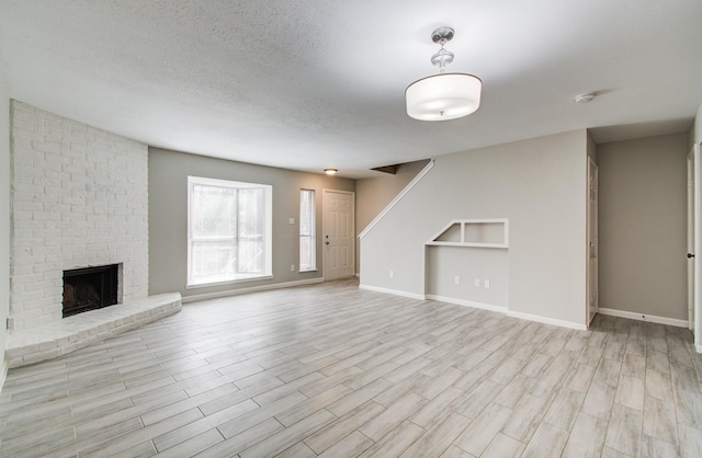 unfurnished living room with a textured ceiling, light wood-style flooring, baseboards, stairs, and a brick fireplace