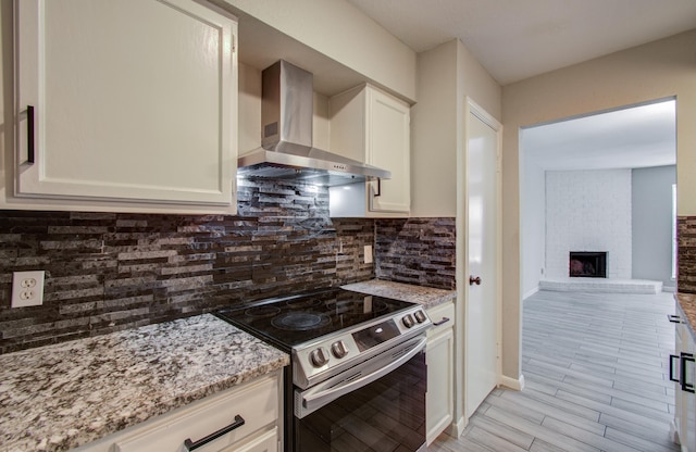 kitchen with tasteful backsplash, electric stove, wall chimney exhaust hood, light stone countertops, and a brick fireplace