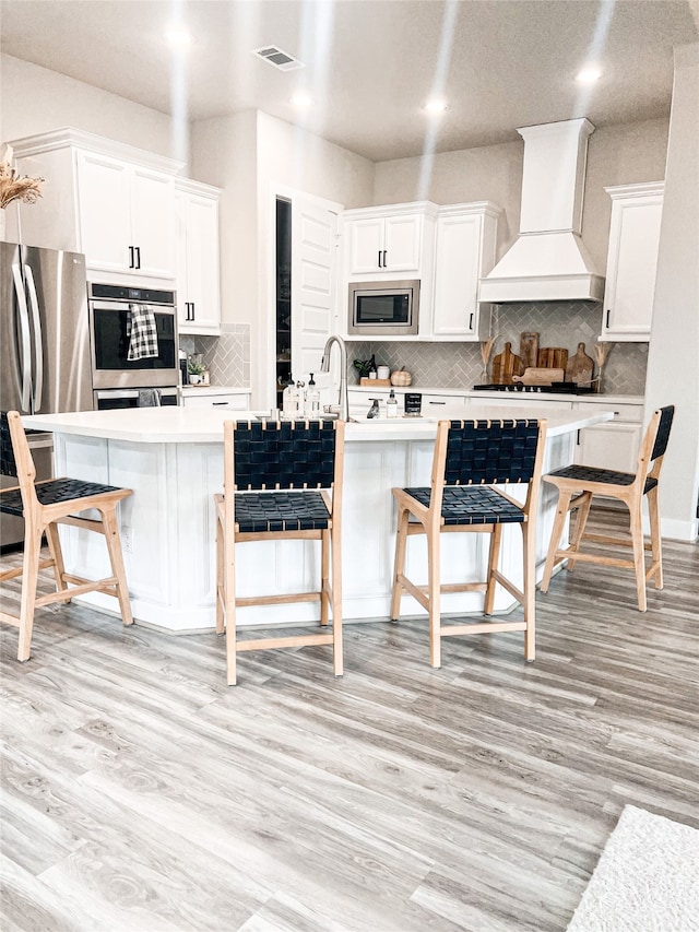 kitchen with appliances with stainless steel finishes, a breakfast bar, custom range hood, white cabinets, and an island with sink