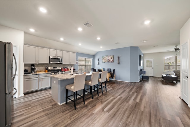 kitchen featuring hardwood / wood-style floors, a kitchen island with sink, ceiling fan, light stone countertops, and appliances with stainless steel finishes