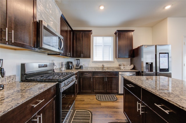 kitchen with appliances with stainless steel finishes, dark brown cabinets, dark hardwood / wood-style floors, light stone countertops, and sink