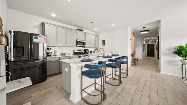 kitchen with stainless steel appliances, a kitchen bar, gray cabinets, an island with sink, and light hardwood / wood-style flooring