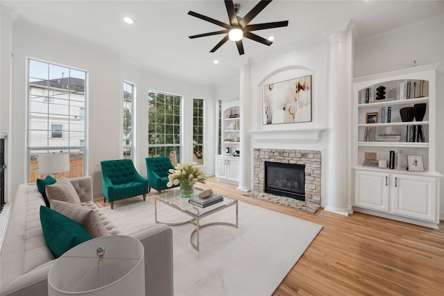 living room featuring built in shelves, ceiling fan, a stone fireplace, crown molding, and light hardwood / wood-style floors