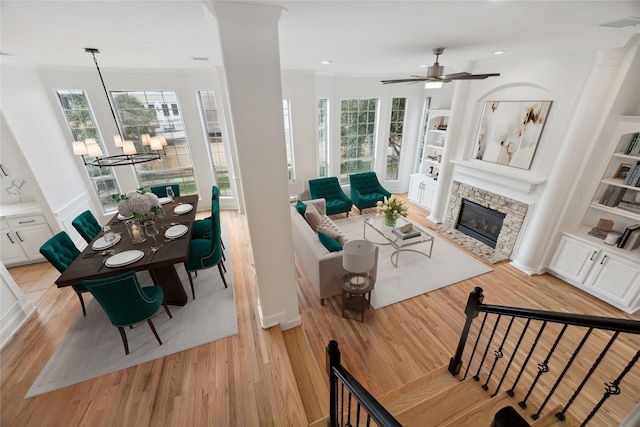 living room featuring built in shelves, ceiling fan, a stone fireplace, crown molding, and light wood-type flooring