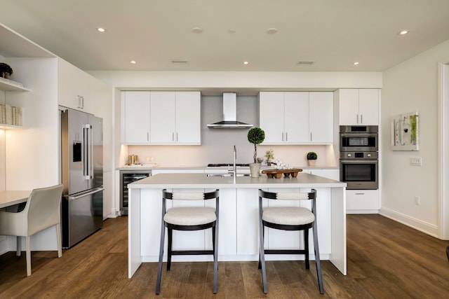 kitchen featuring a center island with sink, white cabinetry, wall chimney range hood, appliances with stainless steel finishes, and beverage cooler