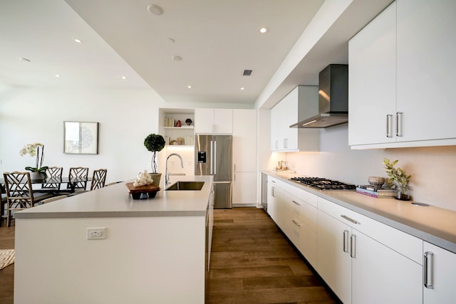 kitchen with dark wood-type flooring, wall chimney exhaust hood, sink, an island with sink, and appliances with stainless steel finishes