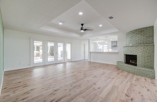 unfurnished living room featuring light hardwood / wood-style floors, ceiling fan, a raised ceiling, a brick fireplace, and french doors