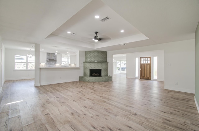 unfurnished living room featuring a brick fireplace, ceiling fan, light wood-type flooring, and a tray ceiling