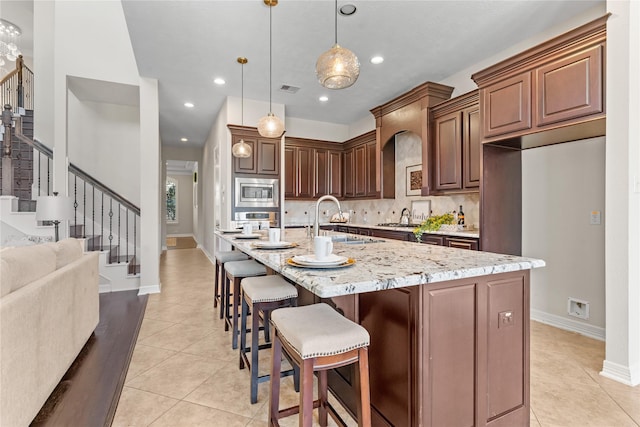 kitchen with light stone countertops, stainless steel microwave, pendant lighting, a kitchen island with sink, and light tile patterned floors